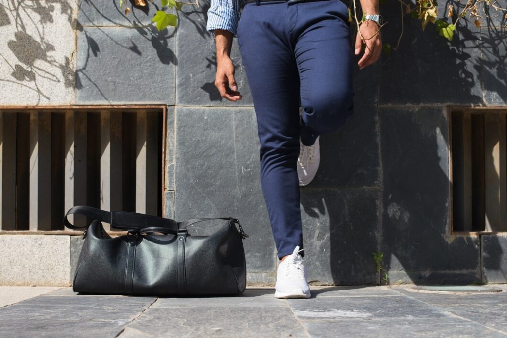 Person in navy pants and white sneakers walking past a black duffel bag on the sidewalk.