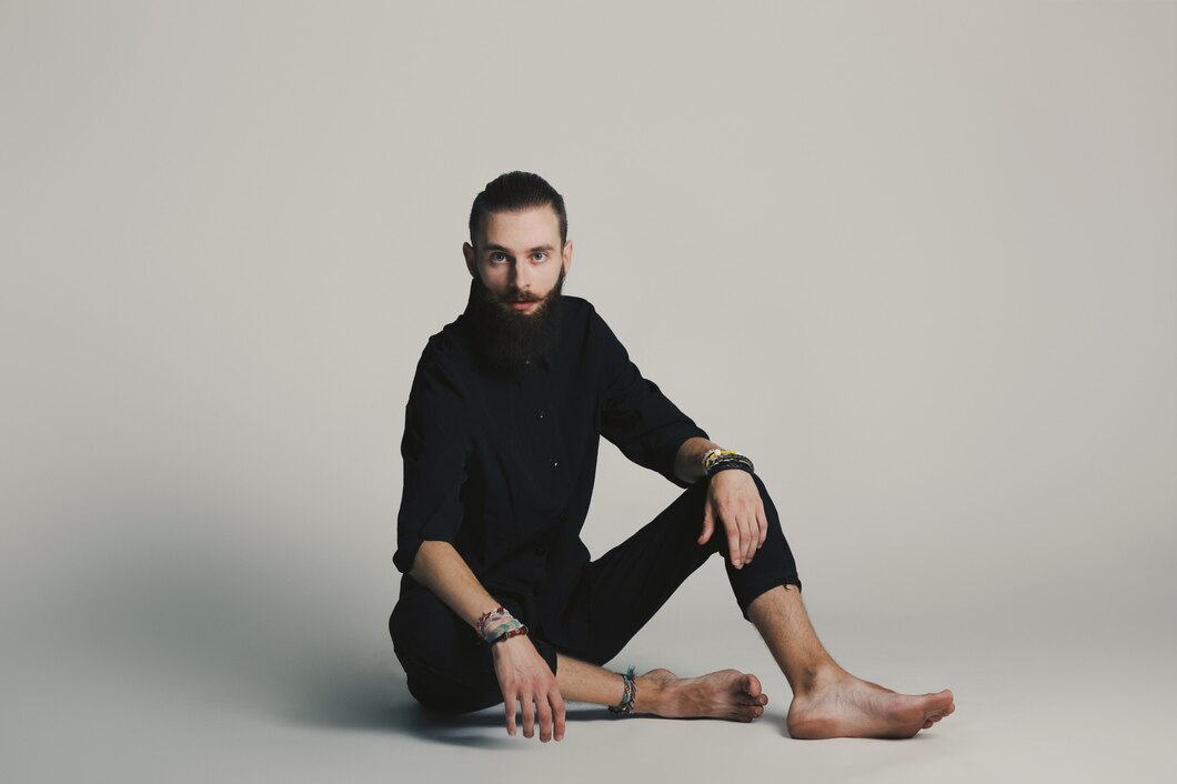 Man with beard sitting cross-legged on the floor in all black, barefoot and relaxed.