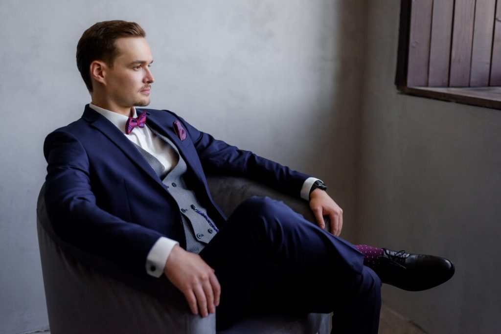 Man in a navy suit with a pink bow tie and vest, seated confidently in a modern chair.