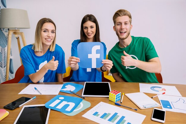 Three young adults sitting at a table holding social media icons, smiling and giving thumbs-up, with charts, tablets, and phones on the desk.