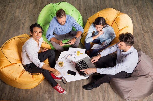 Four colleagues sitting on colorful beanbags around a small table, working together with a laptop, documents, and phones.