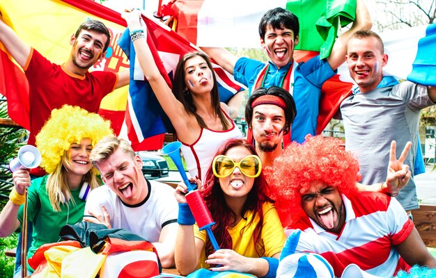 Group of energetic fans with face paint, wigs, and flags cheering and posing enthusiastically outdoors.