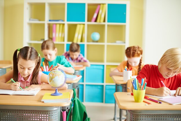 Children focused on writing at desks in a classroom, with bookshelves and supplies neatly arranged in the background.